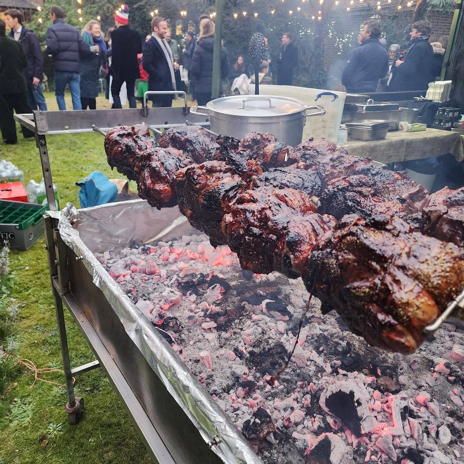 Meat roasting over glowing coals at an evening Christmas party under festoon lights