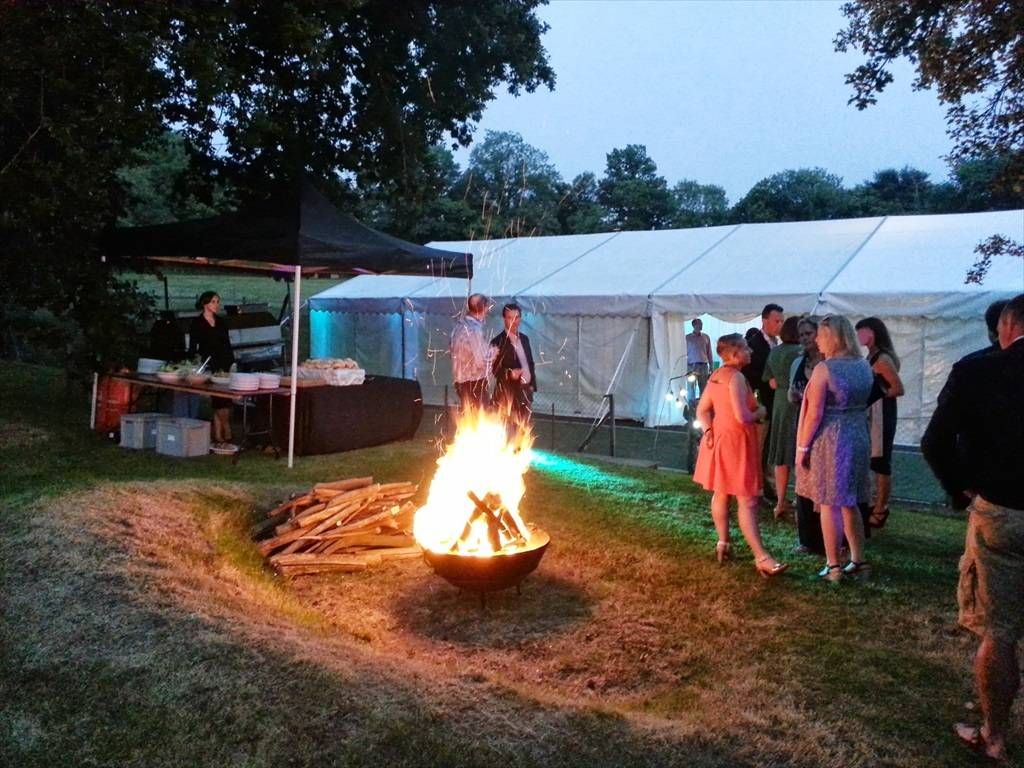 Wedding guests gathered by a fire pit outside a marquee at dusk